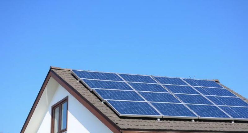 saulės baterijos ant namo stogo saulėtą dieną, simbolizuojančios elektros sąskaitų mažinimą